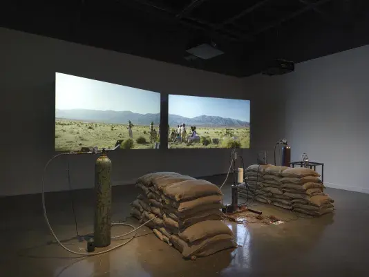 An installation of a rocket test containing sandbags, wiring, and canisters rest in a gallery space in front of two projections depicting people sitting in a desert landscape.
