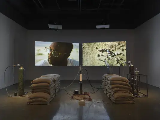 An installation of a rocket test containing sandbags, wiring, and canisters rest in a gallery space in front of two projections depicting people sitting in a desert landscape and a closeup of a man with dark skin tone gazing outwards.