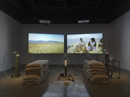 An installation of a rocket test containing sandbags, wiring, and canisters rest in a gallery space in front of two projections depicting people sitting in a desert landscape.