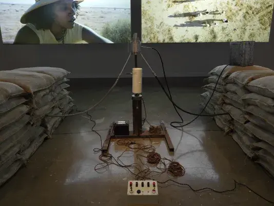 Part of an installation of a rocket test containing sandbags and wiring rest in a gallery space in front of two projections depicting people in a desert landscape.