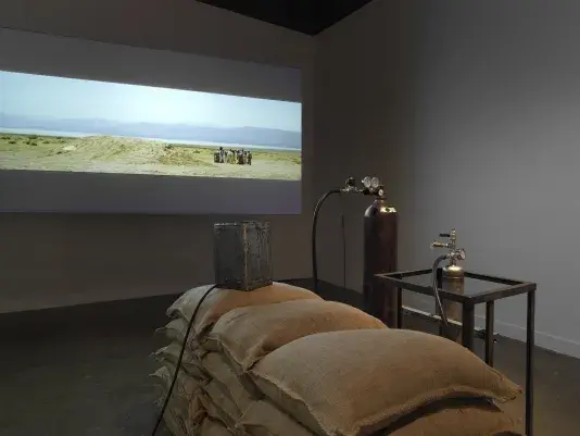 Part of an installation of a rocket test containing sandbags and canisters resting in a gallery space in front of a projection depicting people standing in a desert landscape.
