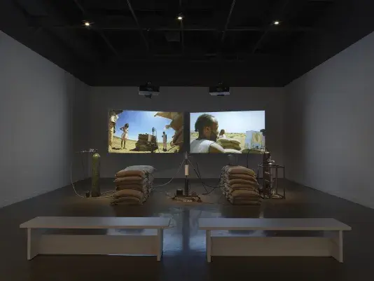 An installation of a rocket test containing sandbags, wiring, and canisters rest in a gallery space in front of two projections depicting people standing in a desert landscape around a similar rocket test set-up. Two white benches sit in the foreground.