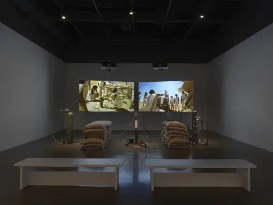 An installation of a rocket test containing sandbags, wiring, and canisters resting in a gallery space in front of two projections depicting people standing in a desert landscape around a similar rocket test set-up. Two white benches sit in the foreground.