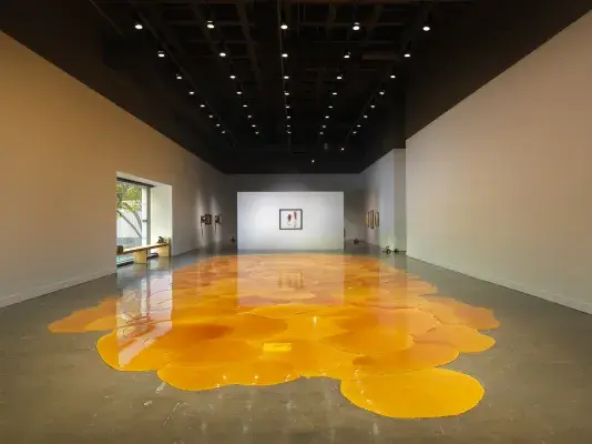 Gallery with an expansive, vibrant orange pool of melted pine rosin that spreads across the floor. Framed artworks hang on the back walls, while a window allows for warm sunlight to flood into the space.