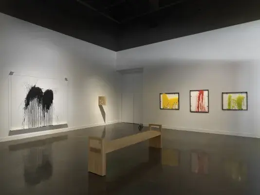 A wooden bench in the middle of a white walled gallery. Three brightly colored abstract artworks in black frames line the wall on the right side, with a large abstract black ink artwork on the adjacent wall to the left.