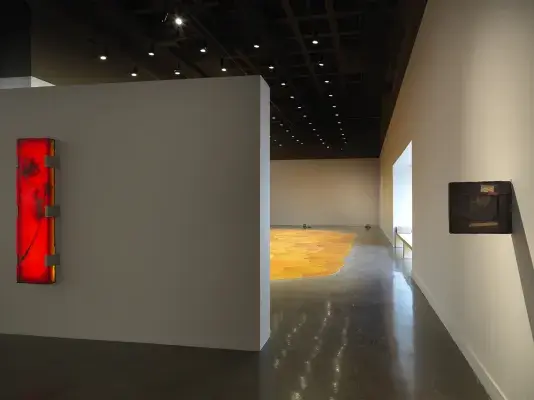 Gallery with an expansive, vibrant orange pool of melted pine rosin that spreads across the floor. Small lego robots are placed throughout the space. A large rectangular illuminated red-orange artwork hangs on a wall on the left side of the frame and a small deep brown framed artwork hangs perpendicular to the wall on the right side.