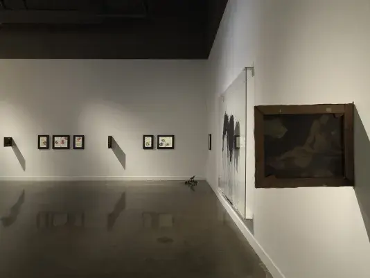 A series of black framed pages of text with colorful botanical ink blots hang in a row on white walls in a dimly lit gallery. A small lego robot sits on the floor in the corner of the space. On the right side of the frame, the artwork closest to the viewer hangs perpendicular to the wall.