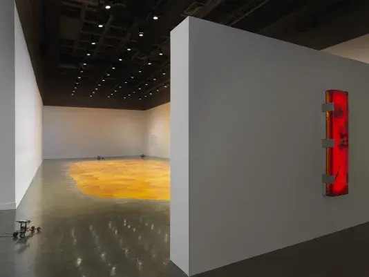 Gallery with an expansive, vibrant orange pool of melted pine rosin that spreads across the floor. Small lego robots are placed throughout the space. A large rectangular illuminated red-orange artwork hangs on a wall on the right side of the frame.
