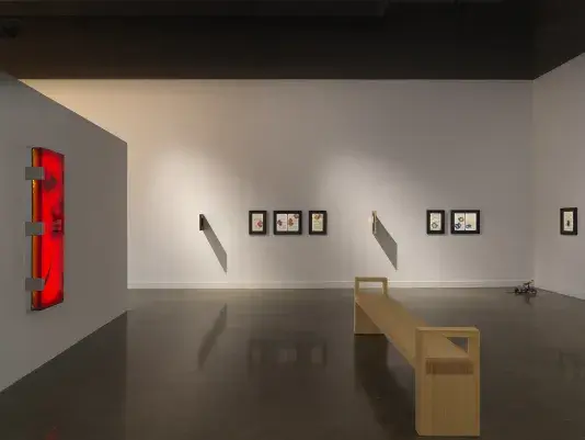 A wooden bench in the middle of a white walled gallery. Small black framed artworks line the back wall. A large rectangular illuminated red-orange artwork hangs on the wall on the left side of the frame.
