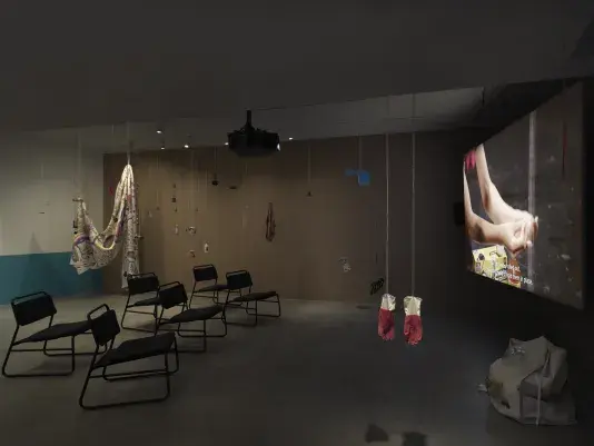 Dark open gallery with a video projected on a large standing screen on the right side of the frame and six black chairs in two rows in the center of the room. A tan canvas bag sits on the floor to the right of the screen. There is a collection of everyday items hanging from strings throughout the space. Two bright red rubber gloves hang in the foreground.
