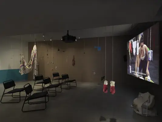 Dark open gallery with a video projected on a large standing screen on the right side of the frame and six black chairs in two rows in the center of the room. A tan canvas bag sits on the floor to the right of the screen. There is a collection of everyday items hanging from strings throughout the space. Two bright red rubber gloves hang in the foreground.