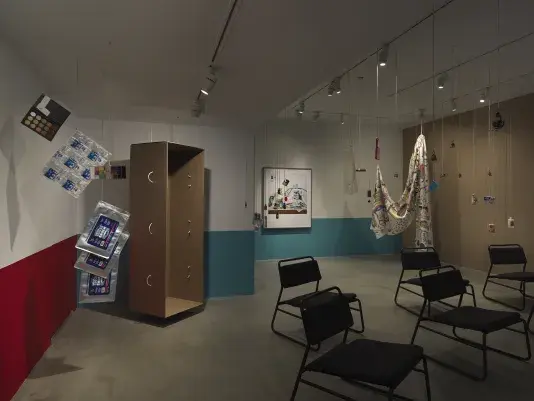 A dimly lit white walled gallery with the bottom half of the wall painted red and text on the left side of the wall. A collection of everyday items hanging from strings throughout the space and a large black framed color photograph hangs on the back wall. A tall, open cardboard structure is suspended in the space and a large printed sheet of fabric hangs on the right side of the frame.