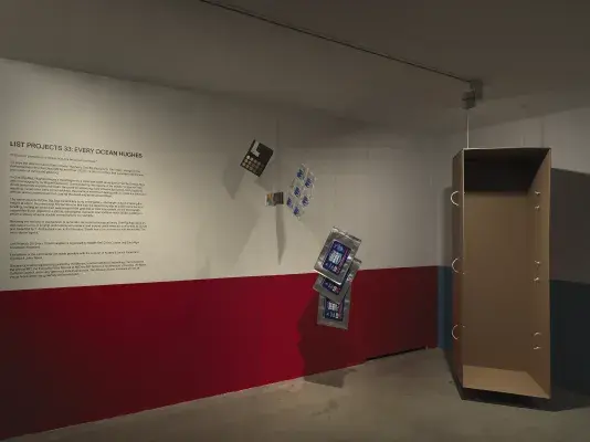 A dimly lit white walled gallery with the bottom half of the wall painted red and text on the left side of the wall. A collection of everyday items hanging from strings throughout the space. A tall, open cardboard structure stands on the right side of the frame.