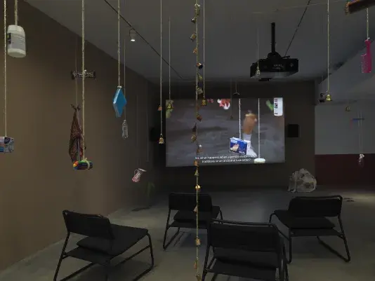 Dark open gallery with a video projected on a large standing screen in the back of the space. There is a collection of everyday items hanging from strings throughout the gallery. A tan canvas bag sits on the floor to the right of the screen and there is a row of chairs in the foreground.