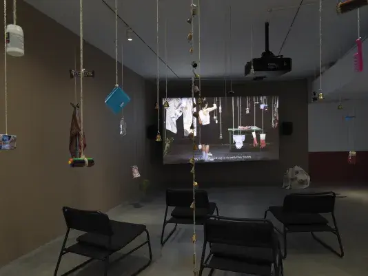 Dark open gallery with a video projected on a large standing screen in the back of the space. There is a collection of everyday items hanging from strings throughout the gallery. A tan canvas bag sits on the floor to the right of the screen and there is a row of chairs in the foreground.