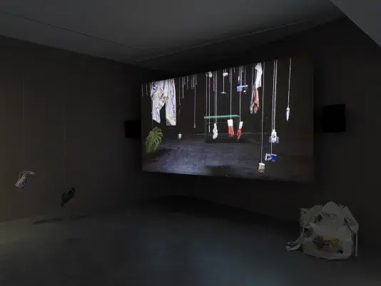 Dark open gallery with a video projected on a large standing screen in the back of the space. There is a collection of everyday items hanging from strings throughout the gallery. A tan canvas bag sits on the floor to the right of the screen.