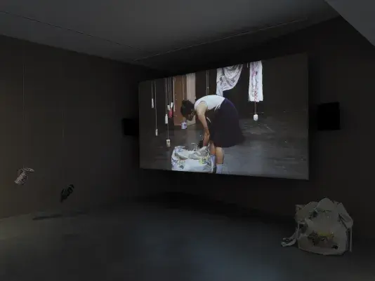 Dark open gallery with a video projected on a large standing screen in the back of the space. There is a collection of everyday items hanging from strings throughout the gallery. A tan canvas bag sits on the floor to the right of the screen.