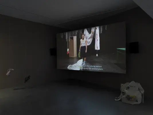 Dark open gallery with a video projected on a large standing screen in the back of the space. There is a collection of everyday items hanging from strings throughout the gallery. A tan canvas bag sits on the floor to the right of the screen.