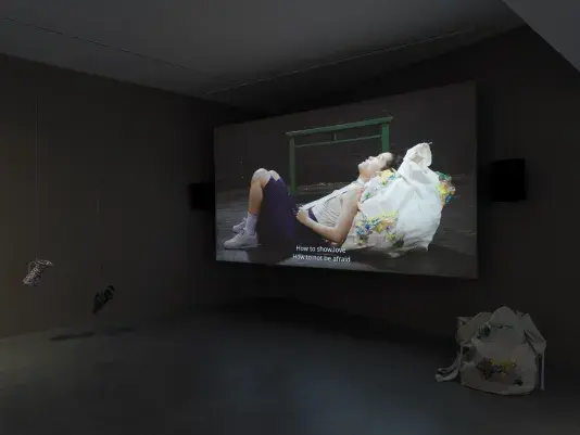 Dark open gallery with a video projected on a large standing screen in the back of the space. There is a collection of everyday items hanging from strings throughout the gallery. A tan canvas bag sits on the floor to the right of the screen.