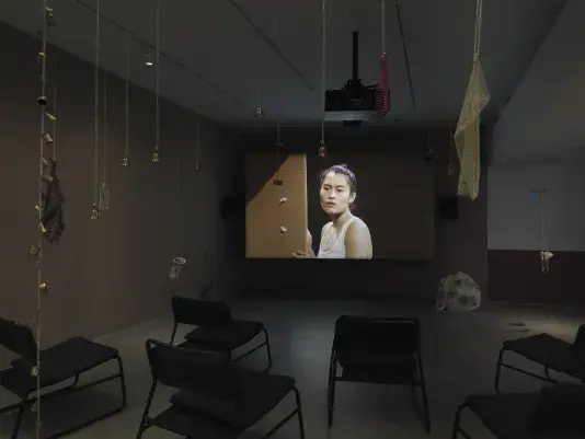 Dark open gallery with a video projected on a large standing screen in the back of the space. There is a collection of everyday items hanging from strings throughout the gallery. A tan canvas bag sits on the floor to the right of the screen and there is a row of chairs in the foreground.