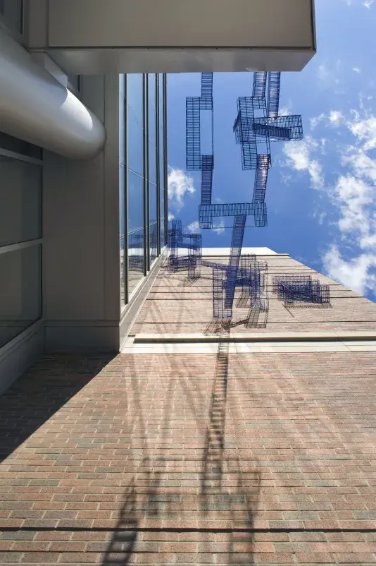Brick wall with bright blue poles resembling a fire escape staircase from underneath looking up at the sculpture and blue sky.
