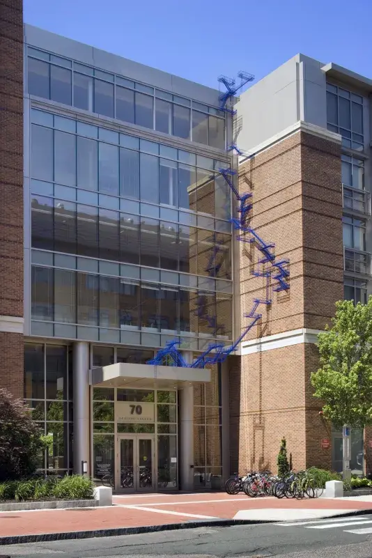 Brick wall with bright blue poles resembling a fire escape staircase is visible on the facade of a brick building with large windows.