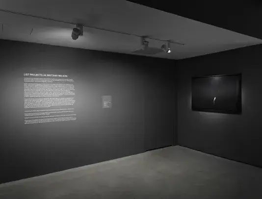 A dimly lit gallery room with dark gray walls features a text panel on the left wall. A framed artwork with a subtle light is on the right wall.