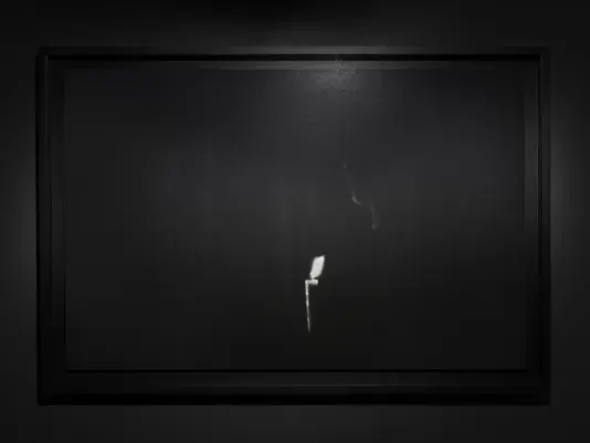 A black-and-white photo of a figure holding a candle in complete darkness. The image is predominantly black, with a lit candle at the bottom center of the frame. A faint silhouette of the figure is partially illuminated by the candlelight.