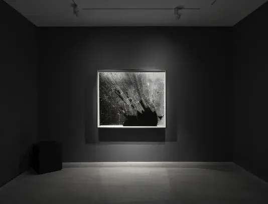 A black-and-white abstract photograph depicting shattered glass is displayed in a dimly lit gallery. The room has dark gray walls, and the photograph is spotlit on the wall.