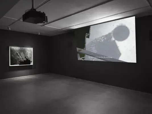 A dark gallery room with two artworks on display. On the left, an abstract black-and-white photograph of shattered glass hangs on a dark gray wall. On the right, a large video projection shows a giant white metal structure.
