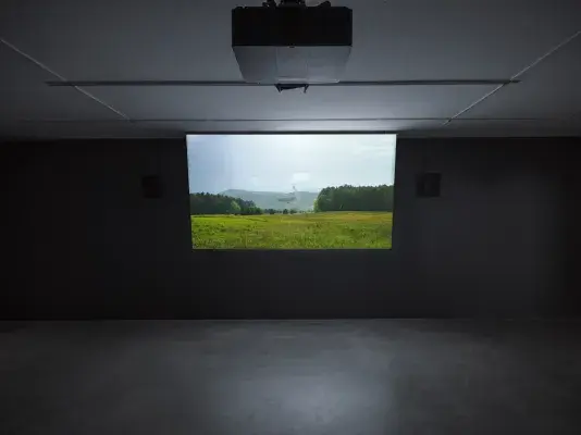 A projector screen in a dark room displays a serene landscape of a green meadow, with mountains and a telescope in the distance.