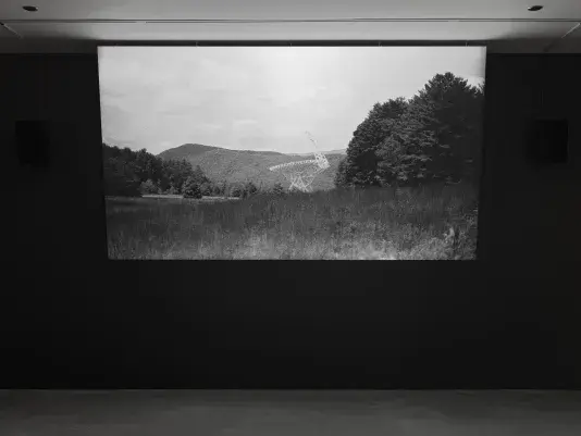 A black and white video is displayed on a screen in a dark room. The video shows a large radio telescope in a field surrounded by dense trees and hills.