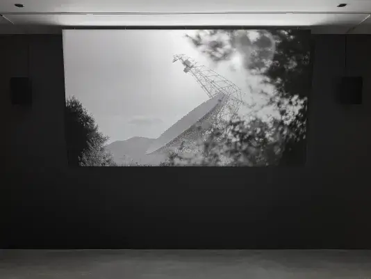 A black-and-white video is displayed on a screen in a dark room. The video shows a large satellite dish against a cloudy sky, partially obscured by trees. The text “in dreams” appears in the video.