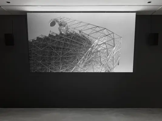A black-and-white video is displayed on a screen in a dark room. The video shows a large, intricate metal lattice structure with complex geometric angles and lines.