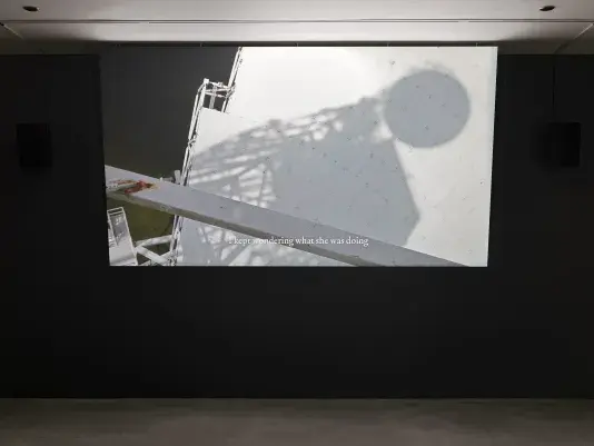 Projected onto a screen on a dark wall is video footage showing the shadow of a circular antenna on a white surface, with the text “I kept wondering what she was doing.” appearing across the bottom of the video.