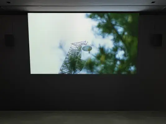 A projected image on a screen in a dark gallery shows a tall metal tower with a dish, surrounded by blurred green foliage.