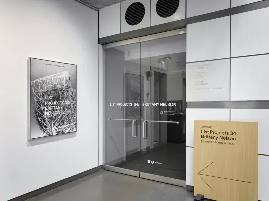 Glass doors display the text “List Projects 34: Brittany Nelson.” A poster hangs on the left wall, featuring the exhibition title over a black-and-white photograph of an intricate metal structure. A wooden sign to the right of the doors lists the exhibition name with an arrow pointing into the gallery.