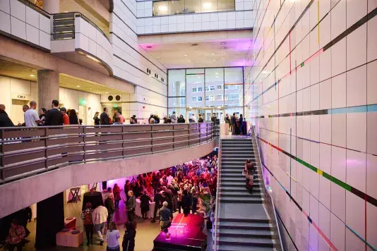 Crowds reside on the first floor and lower level of the List Center. A person is walking up the steps. Colored lights glow throughout the space.