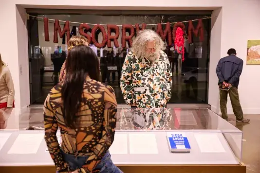 Two visitors look at a vitrine in a gallery. Behind the visitors is a large glass window, and over the window a string of text hangs and reads "I'm Sorry Mom". A man in a blue jacket is reading a wall label in the back.