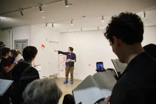 A man with a tie pointing at paint on a gallery wall while an audience watches.