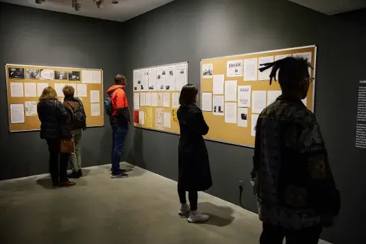 Bulletin boards hang in a dark walled gallery. On the bulletin boards hangs various ephemera. Five gallery visitors face the walls reviewing the content.