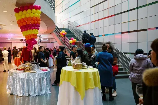A crowd gathers in the List Center's lower atrium. Pink, yellow, and orange balloons in a cone-like structure dnagle from the ceiling over food on a table with a sequin table cloth. Other tables with yellow pink and orange table clothes are scattered throughout the space.