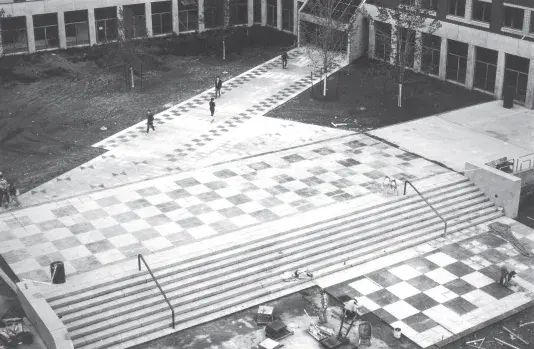 Black and white photograph of a checkered courtyard with greenspace and brick buildings in the backyard.