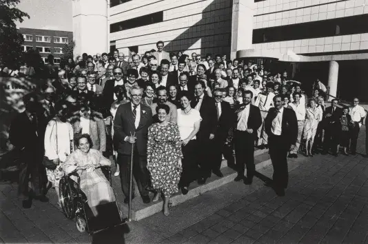 Black and white photograph of a large crowd of people standing on the entrance stairs in front of a building.