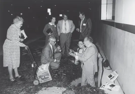 Black and white photograph of seven people gathered in a circle. One man is in a dug up hole in the ground holding a bottle. There are paper booklets on the ground surrounding the hole and one lady is pushing an open box on a hand truck.