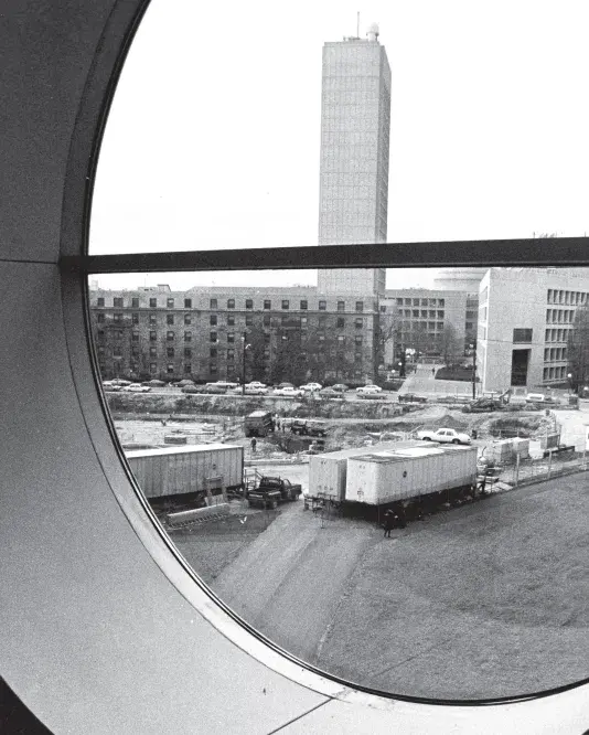 Black and white photograph of a construction site through an oval window. There is a tall rectangular building in the center of the horizon.