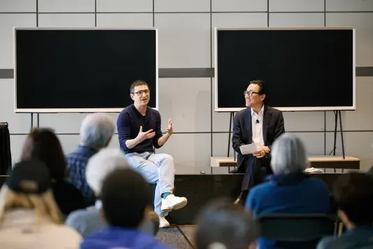 Two people site on a bench in front of two screens speaking to an audience sitting facing them.