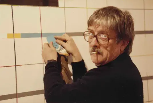 Photograph of a man with blonde hair, large red glasses and a cigarette in his mouth is wearing a black turtleneck. The man is holding up a light blue color swatch to match a color on the wall.