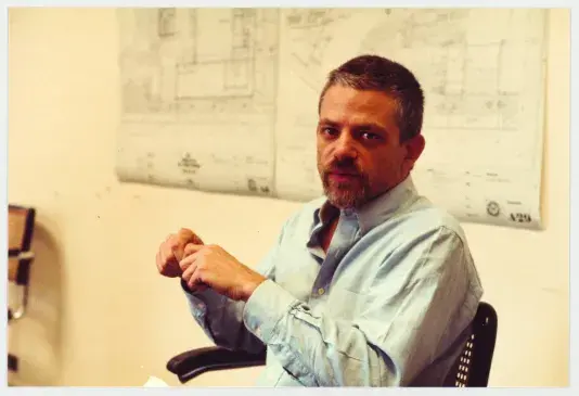 Photograph of a man sitting in an office chair in front of two large black and white building schematics posters hung on the wall wearing a blue button down shirt with his hands together and elbows on the chair rests.
