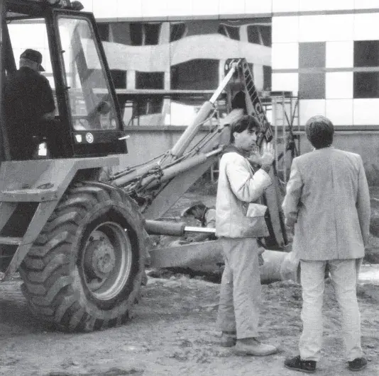 Black and white image of two men in conversation standing in front of a construction backhoe.
